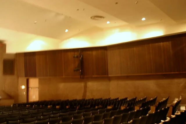 Auditorium seating under a stepped white ceiling with recessed lights and long wood-paneled side walls