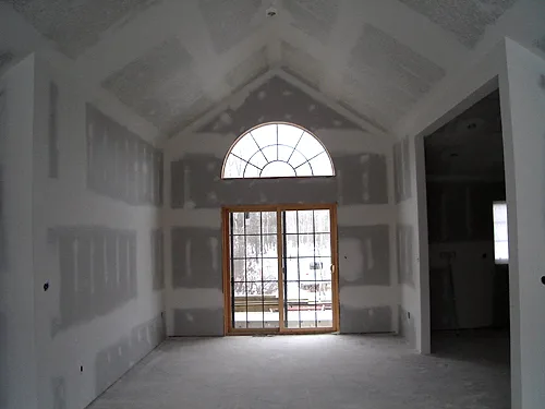 Taped and mudded drywall in a vaulted great room framing an arched window and patio door, before sanding