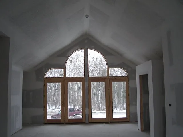 Vaulted great room with taped drywall seams around a wall of tall arched windows looking out on winter woods