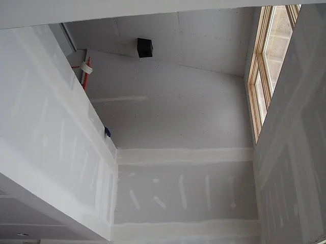 Two-story stairwell with tall drywall walls taped and coated, looking up toward a window and the upper landing