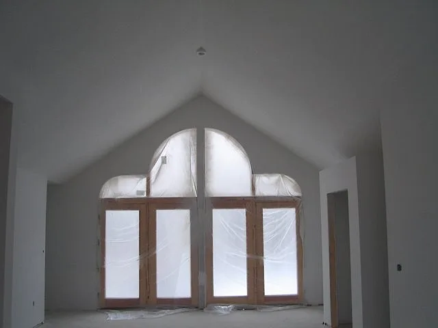 Finished drywall on a tall vaulted wall around arched windows covered in plastic, ready for primer in a new home