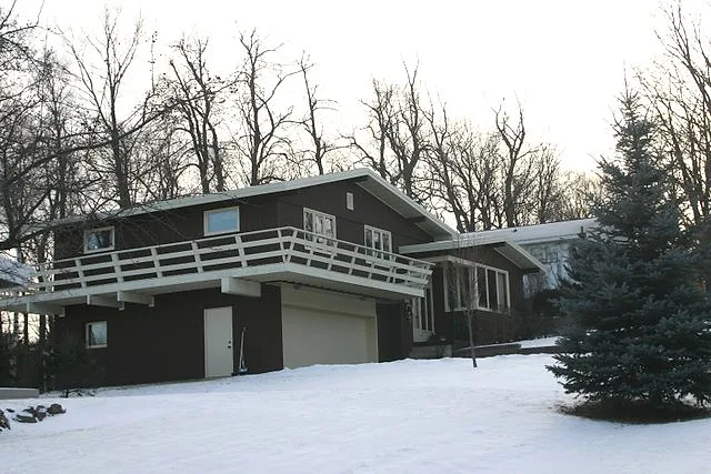 Dark-painted split-level home with white deck railings on a wooded lot, finished exterior paint seen after the first snow