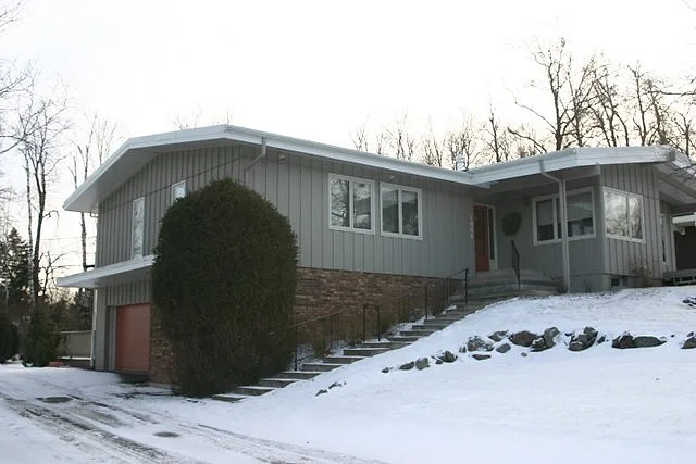 Gray-blue ranch house with white trim and a fieldstone base on a snowy slope, a recent J.A. Houle exterior painting job
