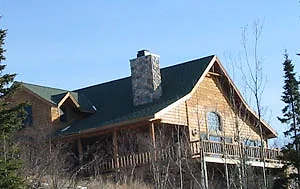 Two-story log home with a green roof and stone chimney after a J.A. Houle exterior stain job, spruce trees behind it