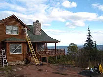 Ladder against a log cabin gable during staining, with a deck, stone chimney and a lake view past the spruce trees